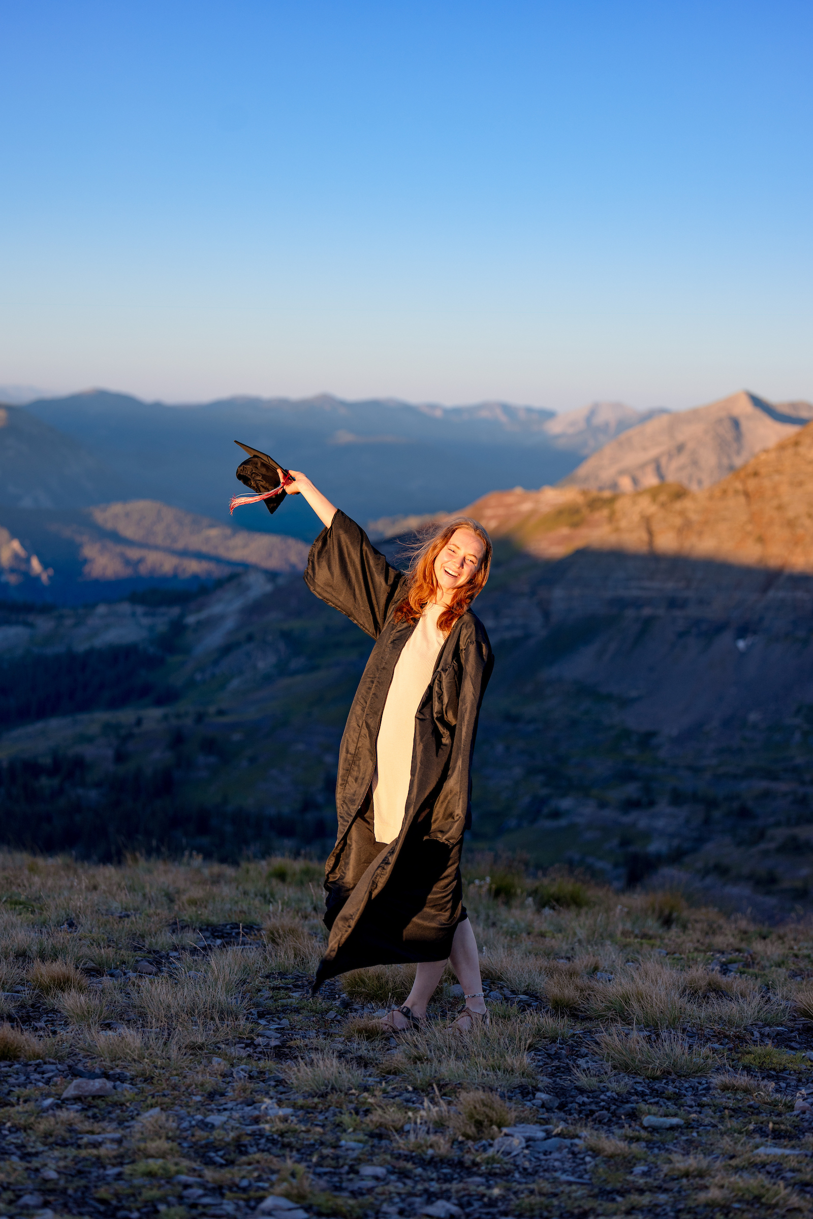 A student wearing a gown and raising her cap stands on a high alpine ridge at sunrise with big mountain views in the background.